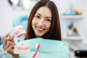 A young woman sitting in a dentist's chair. She is smiling and holding a toothbrush and a model of a set of teeth.